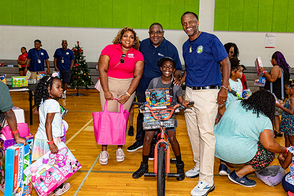 A woman in a red shirt with her child on a bicycle and other toys, standing with Darryl Barnes and Darius Stanton of M-NCPPC.
