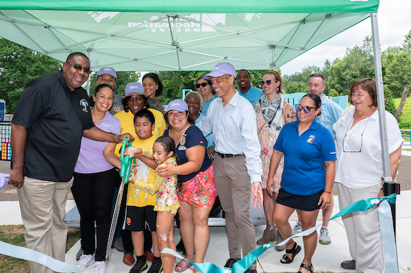 A woman and two children hold giant scissors and cut the ribbon at a ribbon-cutting ceramony. Officials are watching from along side of them.